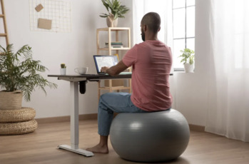 A man in a blue shirt is sitting at his computer desk and stretching his upper body